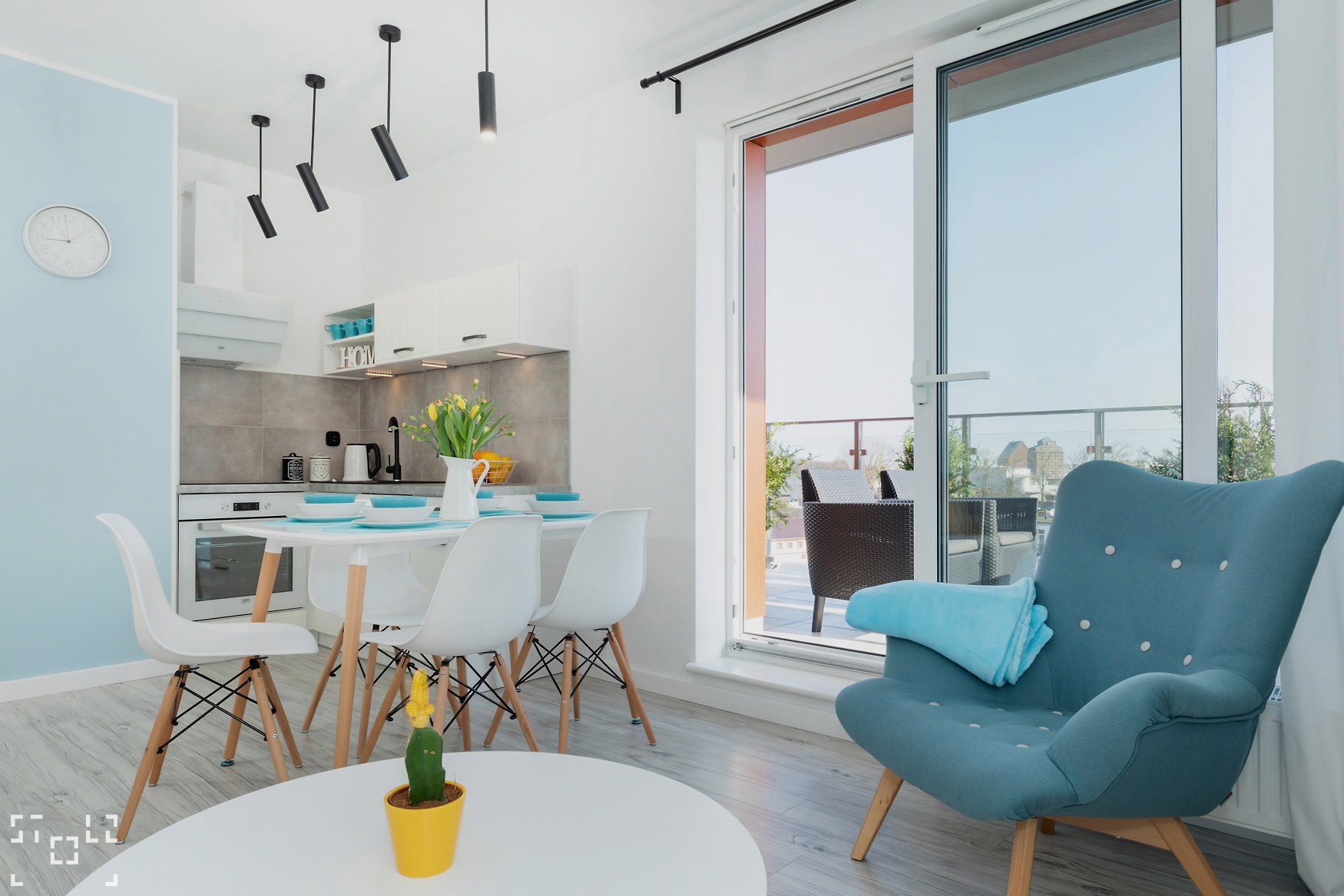 Jasne wnętrze mieszkania z aneksem kuchennym, stołem z krzesłami, balkonem i turkusowym fotelem z kocykiem