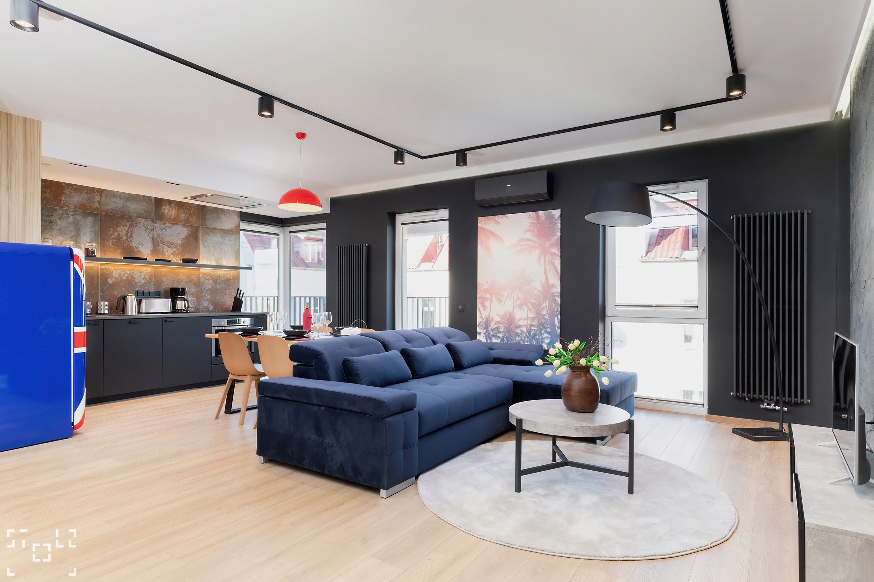 Nowoczesne wnętrze mieszkania z granatową sofą narożną, okrągłym dywanem, stolikiem kawowym, kuchnią w tle i lodówką w barwach flagi brytyjskiej.