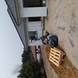 Adam wysota - Pracownik na drabinie maluje elewację domu jednorodzinnego, widoczne czarne worki na śmieci i paleta drewniana.