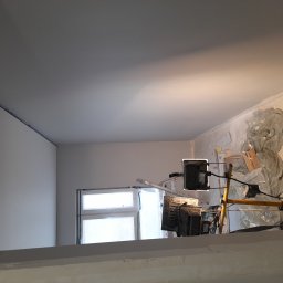 Adam wysota - Wnętrze w trakcie prac: świeżo pomalowany sufit i ściany w odcieniach szarości, zabezpieczone okno folią, widoczne lampy budowlane i drabina.