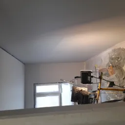 Wnętrze w trakcie prac: świeżo pomalowany sufit i ściany w odcieniach szarości, zabezpieczone okno folią, widoczne lampy budowlane i drabina.