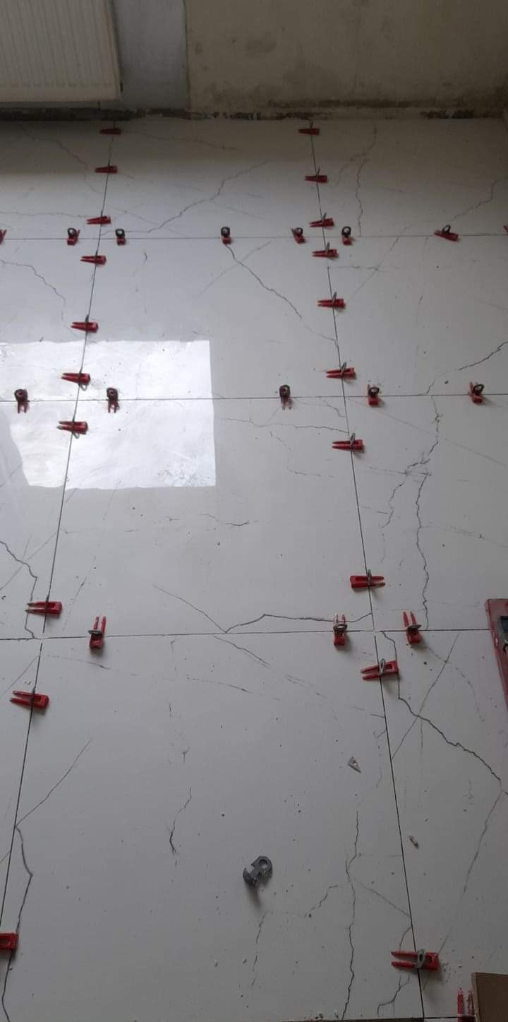 Układanie płytek imitujących marmur z użyciem systemu poziomowania płytek z czerwonymi klipsami i klinami, widoczny fragment ściany w stanie surowym.