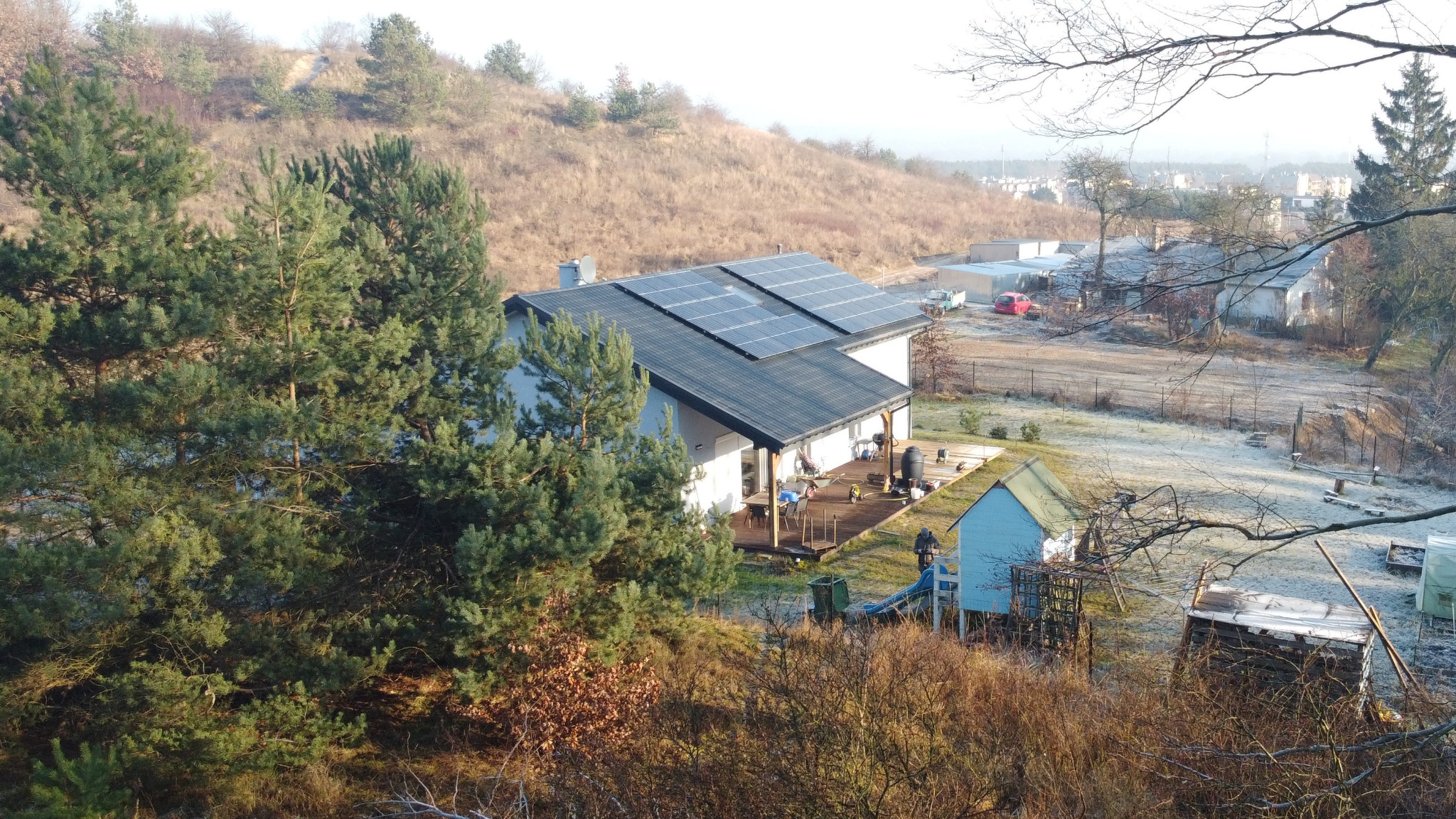 Dom jednorodzinny z panelami słonecznymi na dachu, widok z góry na posesję w jesiennej scenerii z drzewami iglastymi i lekkim oszronieniem.