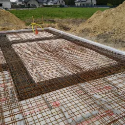 Zbrojenie fundamentów domu jednorodzinnego, widoczne stalowe pręty tworzące siatkę, pod spodem biała izolacja, w tle zabudowa mieszkalna i hałda ziemi.
