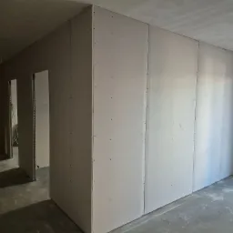 Wnętrze w trakcie prac: ściany działowe z płyt g-k, widoczne łączenia i metalowe profile przy otworach drzwiowych, surowy beton na podłodze.