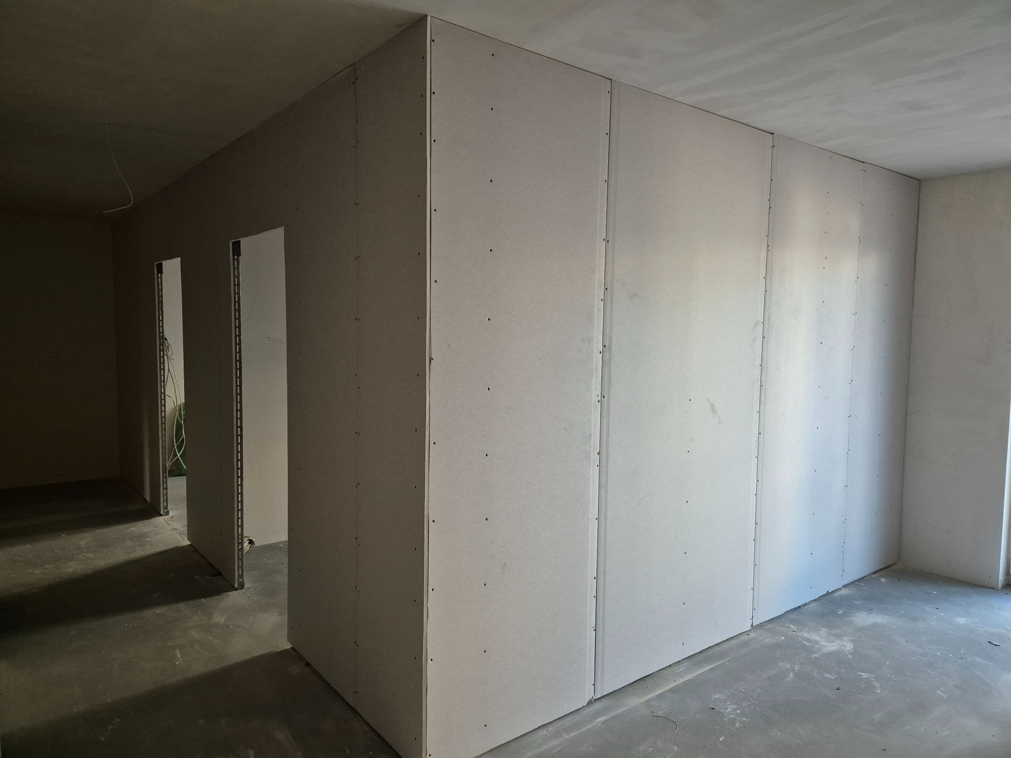 Wnętrze w trakcie prac: ściany działowe z płyt g-k, widoczne łączenia i metalowe profile przy otworach drzwiowych, surowy beton na podłodze.