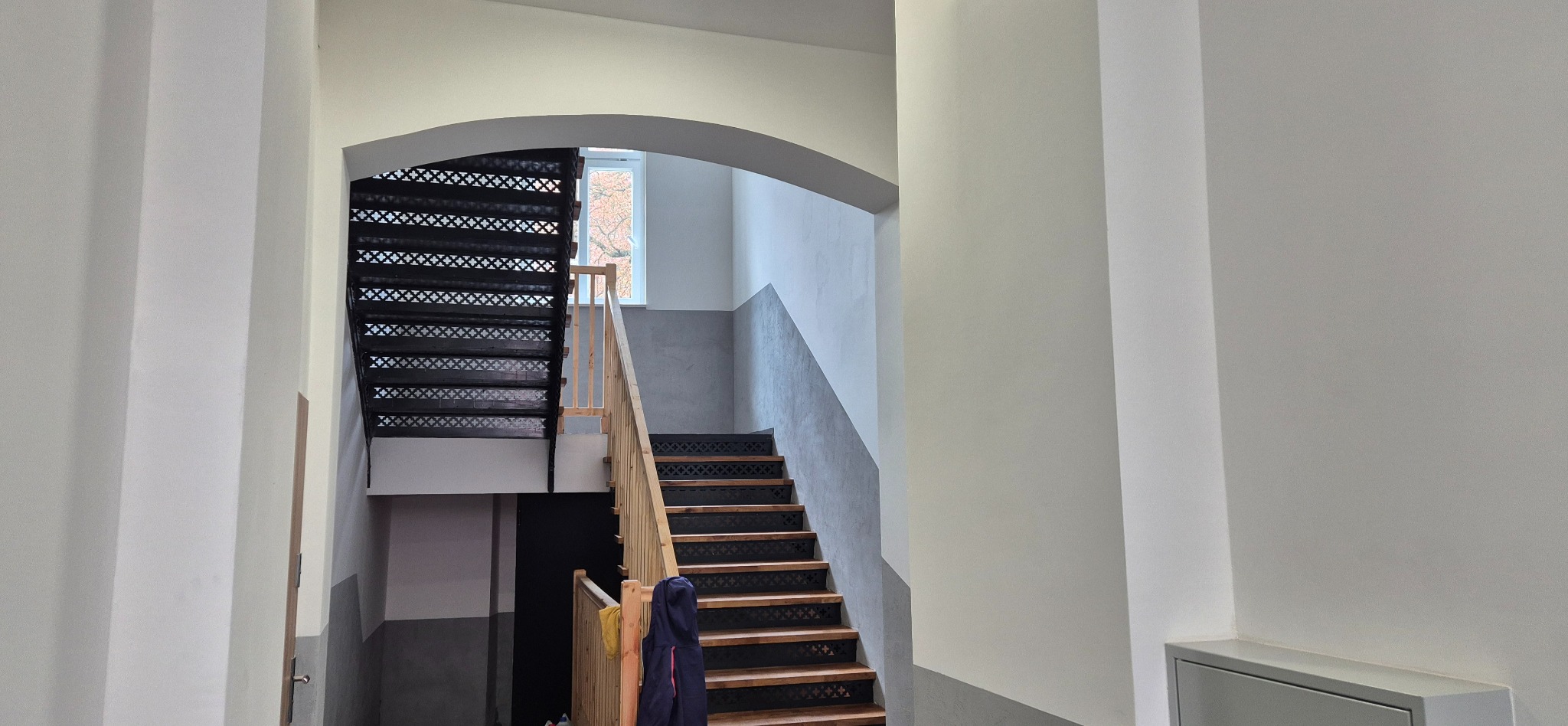 Odnowiona klatka schodowa w kamienicy z drewnianymi schodami i metalową balustradą. Ściany pomalowane w tonacji szaro-białej. Widoczne okno na końcu klatki.