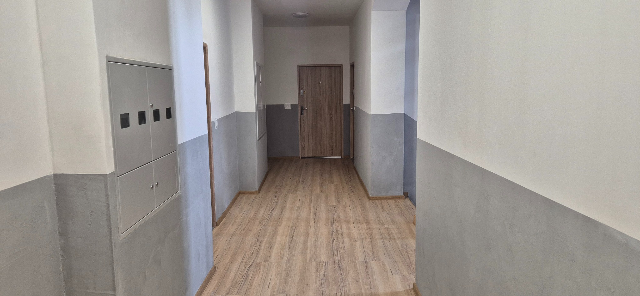 Korytarz po remoncie: panele podłogowe, szare ściany do połowy wysokości, białe powyżej, drzwi wejściowe, szafka elektryczna. Widok w perspektywie.