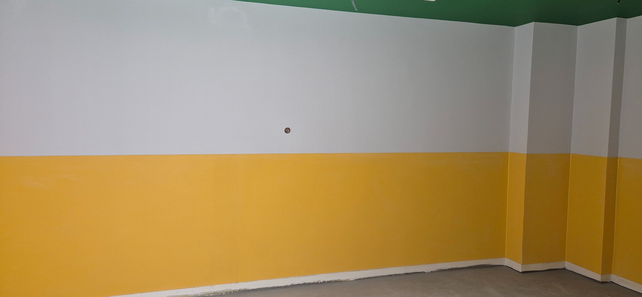 Ściana po malowaniu: góra biała, dół żółty, zielony sufit. Widoczny fragment podłogi i narożnik. Realizacja w pomieszczeniu oświetlonym naturalnym światłem.