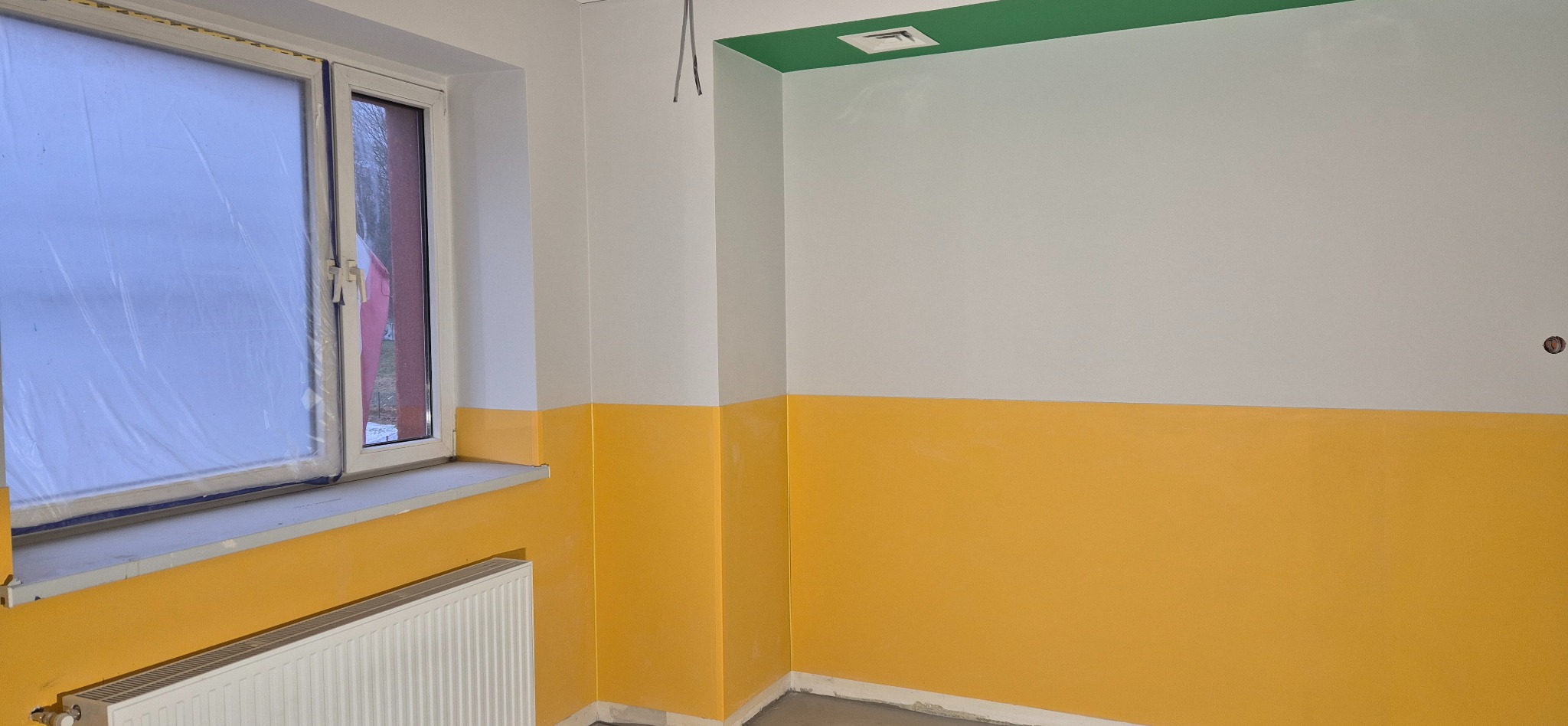 Wnętrze po malowaniu: ściany dwukolorowe, biało-żółte, z zielonym akcentem na suficie. Okno zabezpieczone folią, widoczny grzejnik. Adaptacja budynku klubowego.