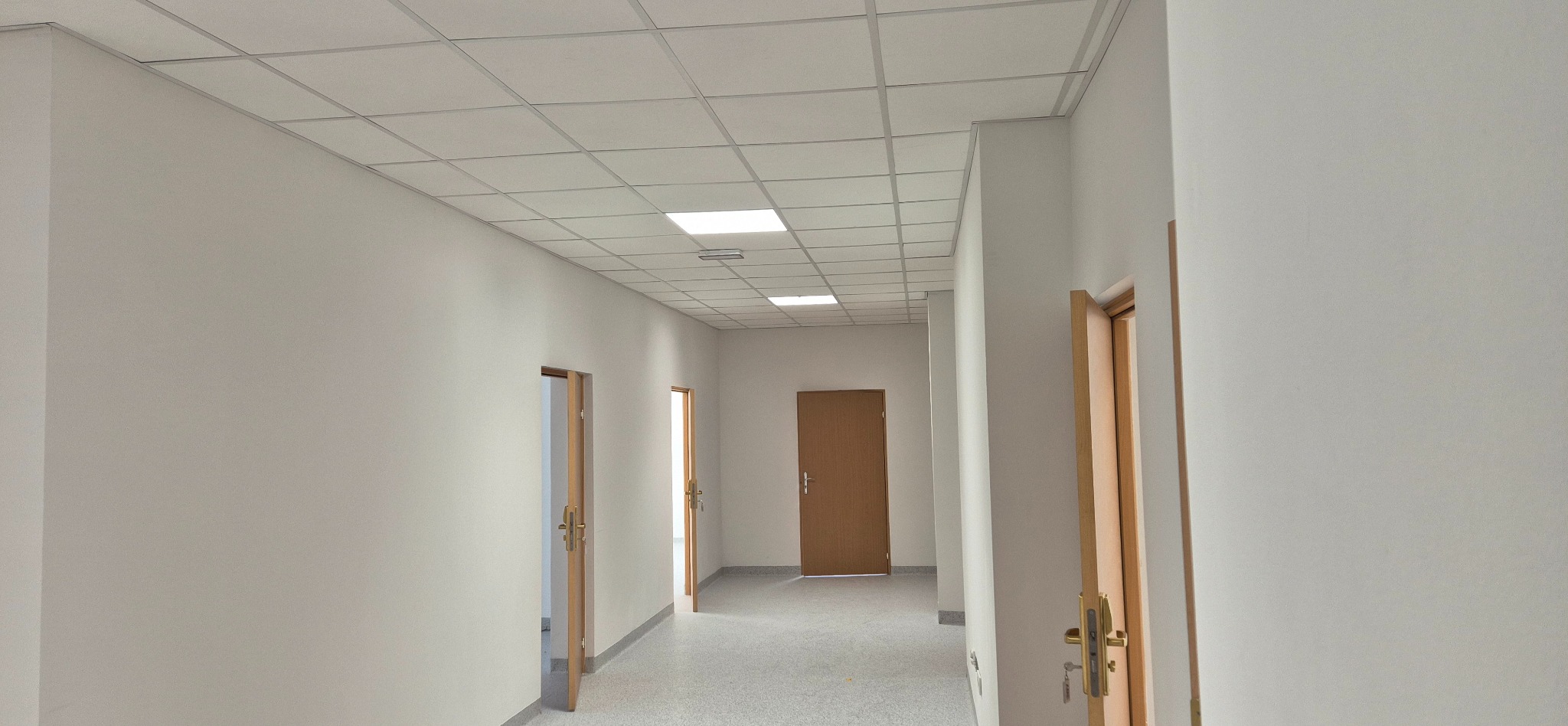 Jasny korytarz biurowy po remoncie. Widoczne świeżo pomalowane ściany, nowe drzwi i sufit z kasetonami. Czyste, minimalistyczne wykończenie wnętrza.