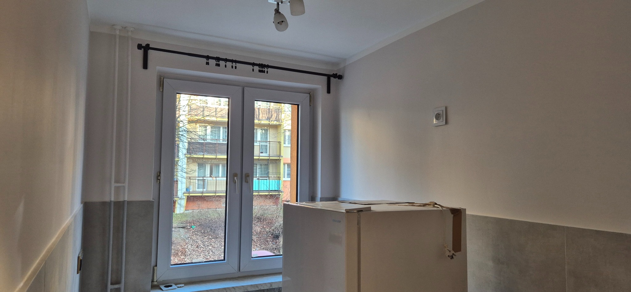 Wnętrze pokoju po remoncie: świeżo pomalowane ściany w odcieniach bieli, okno z czarnym karniszem, widok na blok z balkonami. Częściowo widoczna lodówka.