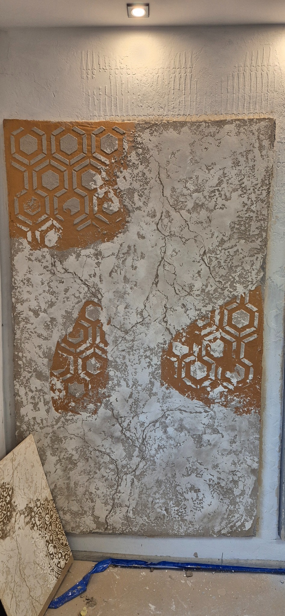 Dekoracyjny tynk z geometrycznym wzorem w odcieniach beżu i szarości, imitujący spękania. Widoczne fragmenty podłoża i niebieska taśma malarska.