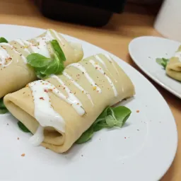 Dwa naleśniki zawinięte w rulony, polane białym sosem i posypane czerwoną papryką, udekorowane zielonymi listkami sałaty, podane na białym talerzu na drewnianym stole.