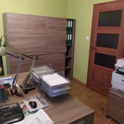 Biuro z drewnianym biurkiem, na którym stoi komputer, lampka, kwiaty w wazonie, kalkulator i stosy dokumentów w metalowych tackach. W tle szafa z segregatorami i drukarka obok drewnianych drzwi.