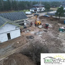Odwierty pod gruntową pompę ciepła na budowie domu jednorodzinnego w Trzcianach, widok z góry na żółtą wiertnicę, mini koparkę i fundamenty budynku z częściowo położonym dachem.