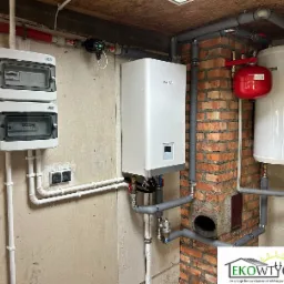 Kompaktowa instalacja grzewcza Kaisai 10kW w kotłowni z widoczną pompą ciepła, zbiornikiem buforowym, naczyniem przeponowym i rozdzielnicą elektryczną na ścianie z surowego betonu.