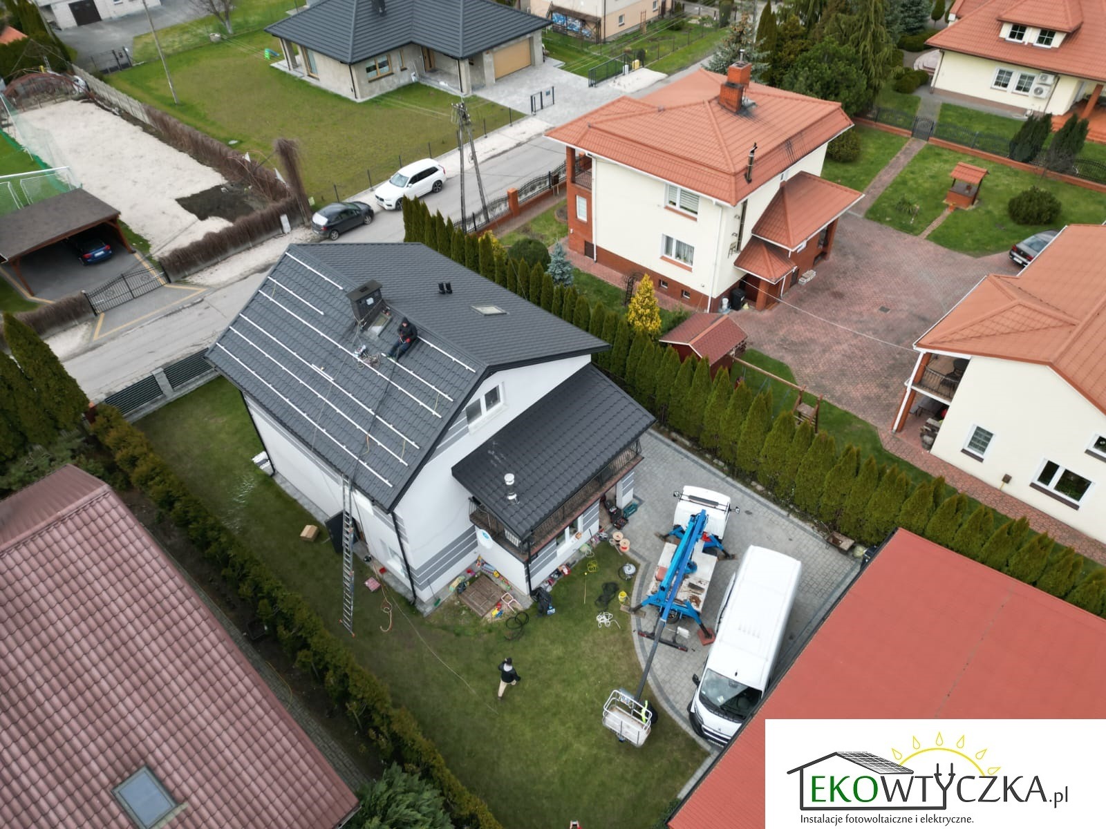 Widok z góry na dom z instalacją paneli słonecznych na dachu, dwóch instalatorów pracuje na dachu, podnośnik koszowy i samochód dostawczy zaparkowane obok, trawnik i żywopłot otaczają budynek.