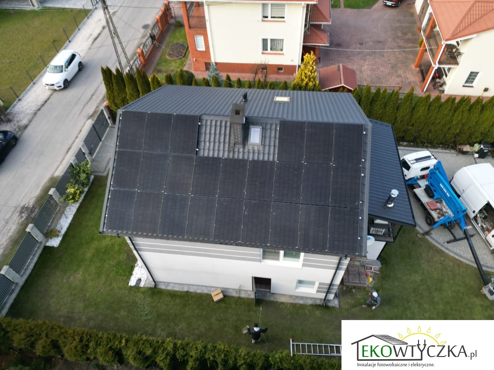 Widok z góry na dom z częściowo zamontowanymi czarnymi panelami słonecznymi na dachu, obok komina i okna dachowego; na trawniku pracownik i drabina, w tle podnośnik koszowy i samochody.