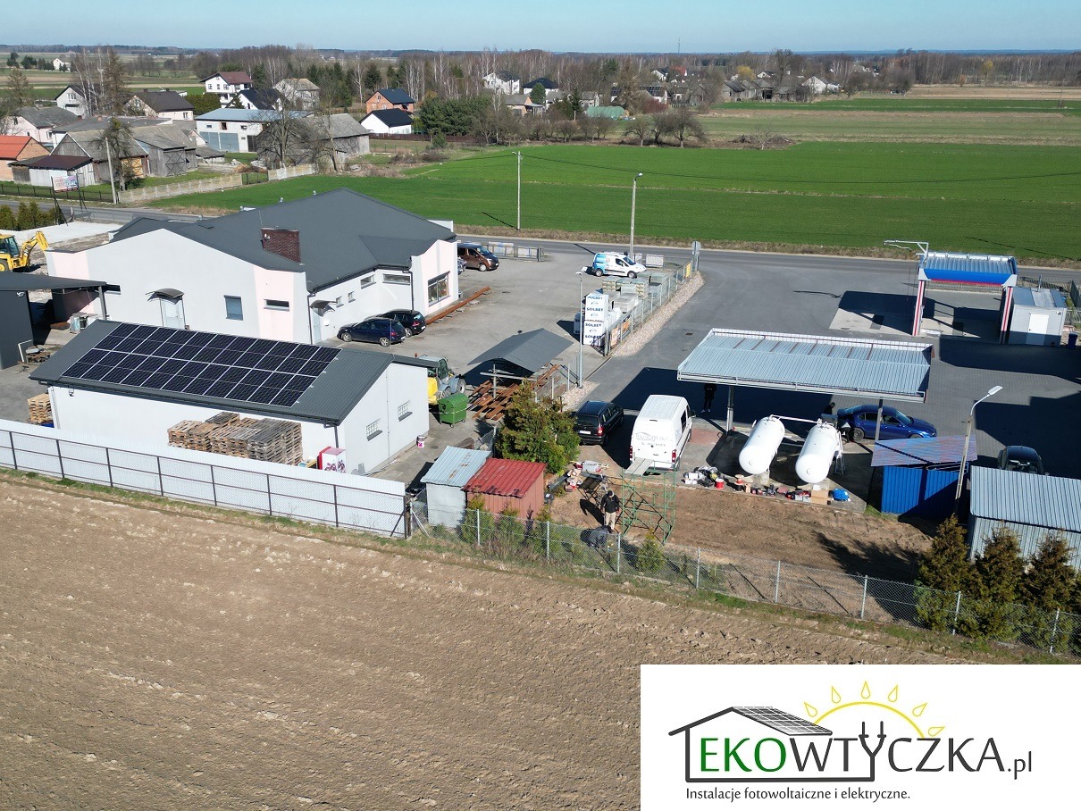 Widok z góry na budynek z panelami słonecznymi na dachu, teren firmy z zaparkowanymi samochodami i zbiornikami, w tle zabudowa wiejska i pola uprawne.