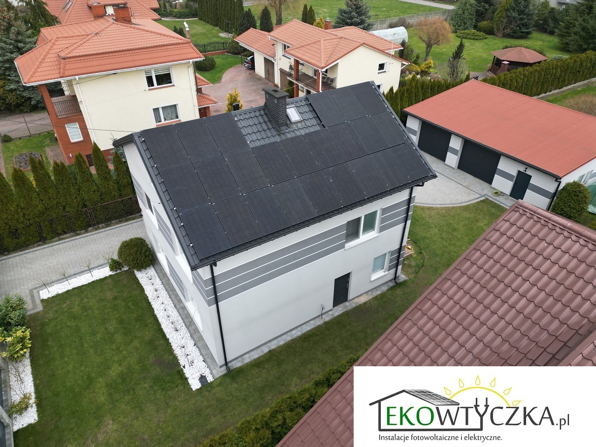 Widok z góry na dom jednorodzinny z panelami słonecznymi na dachu, otoczony zielonym trawnikiem i białymi kamieniami ozdobnymi, w tle inne domy z czerwonymi dachami.