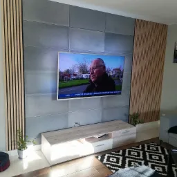 Ściana telewizyjna z szarych paneli betonowych i drewnianych listew pionowych, podświetlany stolik RTV, brązowa podłoga imitująca drewno, czarno-biały dywan geometryczny, roślina doniczkowa po...