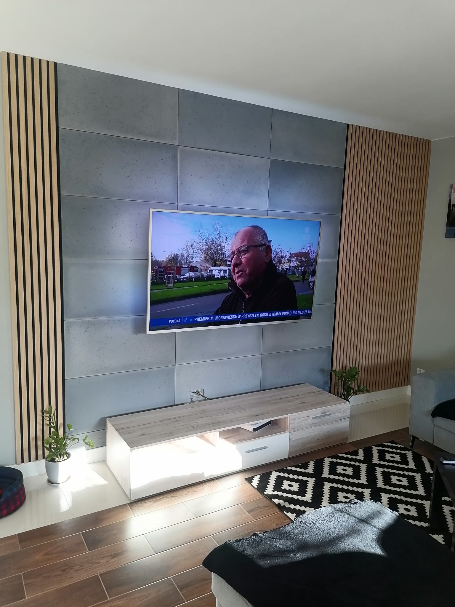 Ściana telewizyjna z szarych paneli betonowych i drewnianych listew pionowych, podświetlany stolik RTV, brązowa podłoga imitująca drewno, czarno-biały dywan geometryczny, roślina doniczkowa po...