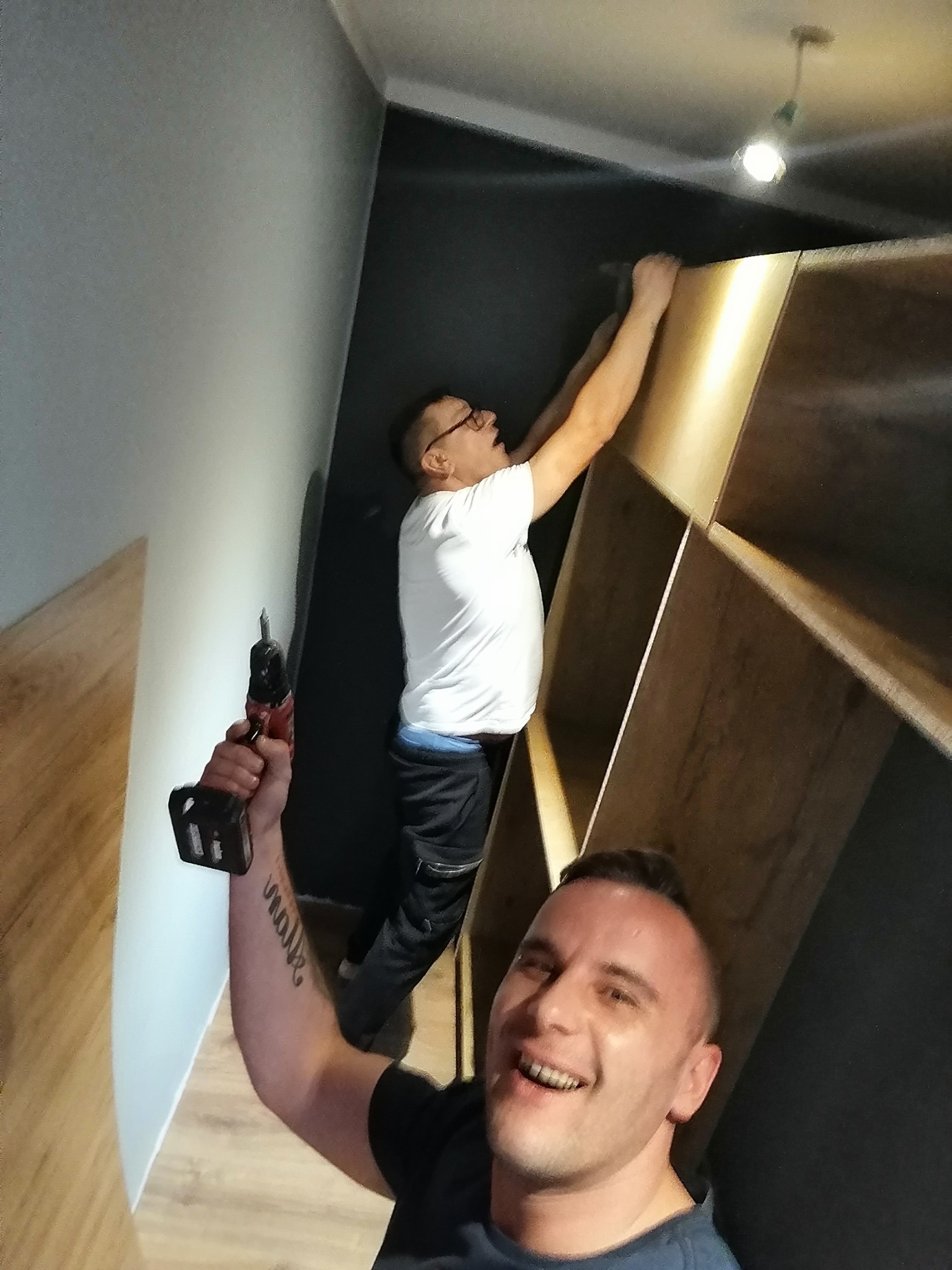 Dwóch mężczyzn montuje drewniany regał w ciemnym pomieszczeniu, jeden z nich uśmiecha się trzymając wiertarkę, drugi przytrzymuje półkę pod sufitem.