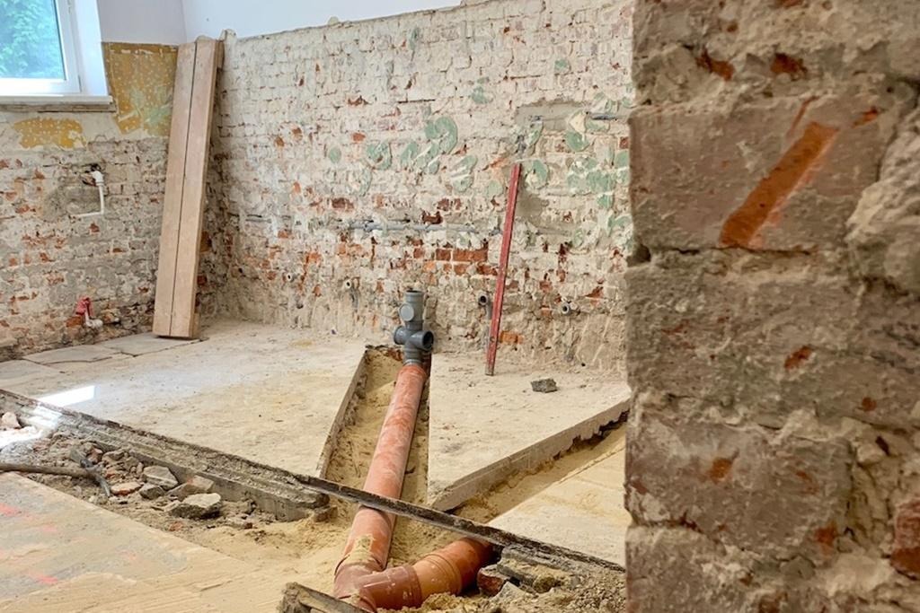 Remont łazienki w starym budynku: odsłonięta cegła na ścianach z widocznymi instalacjami hydraulicznymi, nowe rury kanalizacyjne w wykopanych bruzdach w podłodze, poziomica oparta o ścianę.