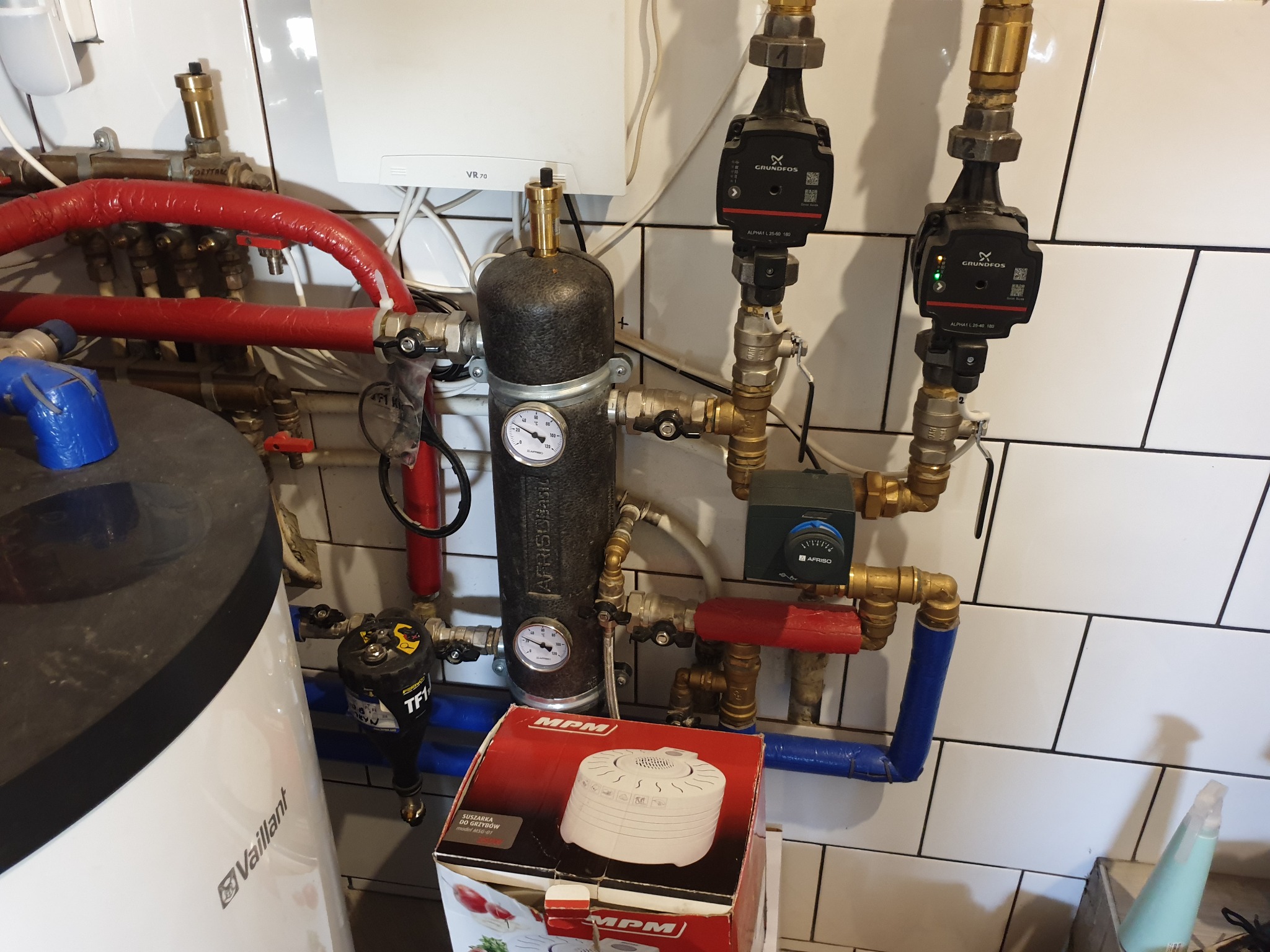 Instalacja centralnego ogrzewania z kotłem Vaillant, pompami obiegowymi Grundfos i zabezpieczeniem Afriso, na tle białych płytek. Na pierwszym planie pudełko suszarki do grzybów MPM.