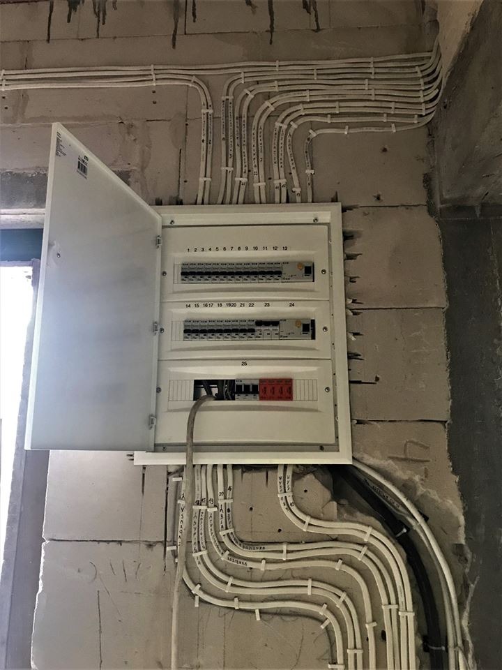 Otwarta skrzynka rozdzielcza z numerowanymi bezpiecznikami na surowej ścianie z betonu komórkowego, z widocznymi przewodami elektrycznymi przymocowanymi do ściany opaskami zaciskowymi.