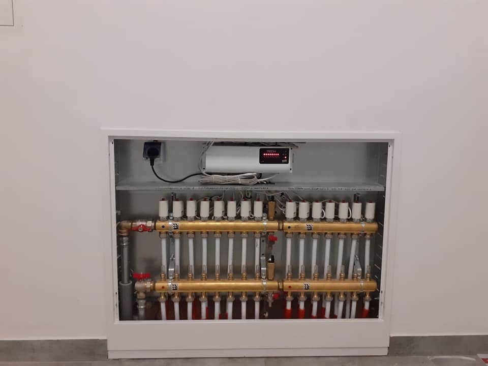 Rozdzielacz ogrzewania podłogowego w białej obudowie, widoczne rury, zawory i sterownik TECH z wyświetlaczem.