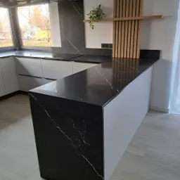 Nowoczesna kuchnia z czarnym blatem z marmurkowym wzorem, białymi szafkami i drewnianymi elementami dekoracyjnymi na ścianie.