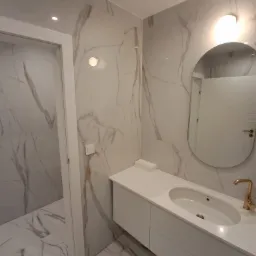 Nowoczesna łazienka z białymi marmurowymi płytkami na ścianach i podłodze, białą szafką pod umywalką z blatem i złotą baterią oraz owalnym lustrem z podświetleniem