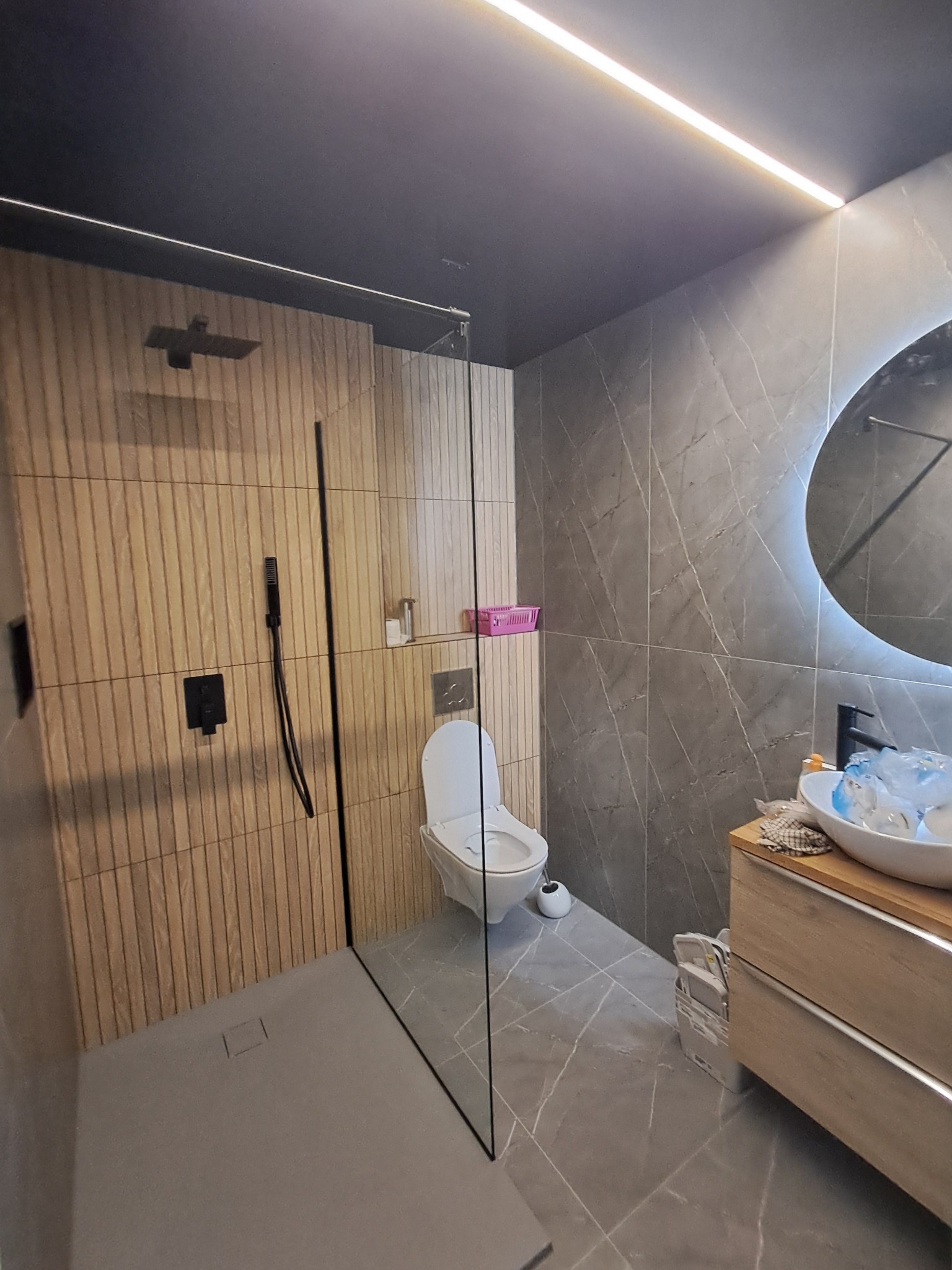 Nowoczesna łazienka z kabiną prysznicową typu walk-in, drewnopodobnymi płytkami ściennymi w strefie prysznica, szarą marmurową okładziną na pozostałych ścianach, podświetlanym lustrem i wiszącą...