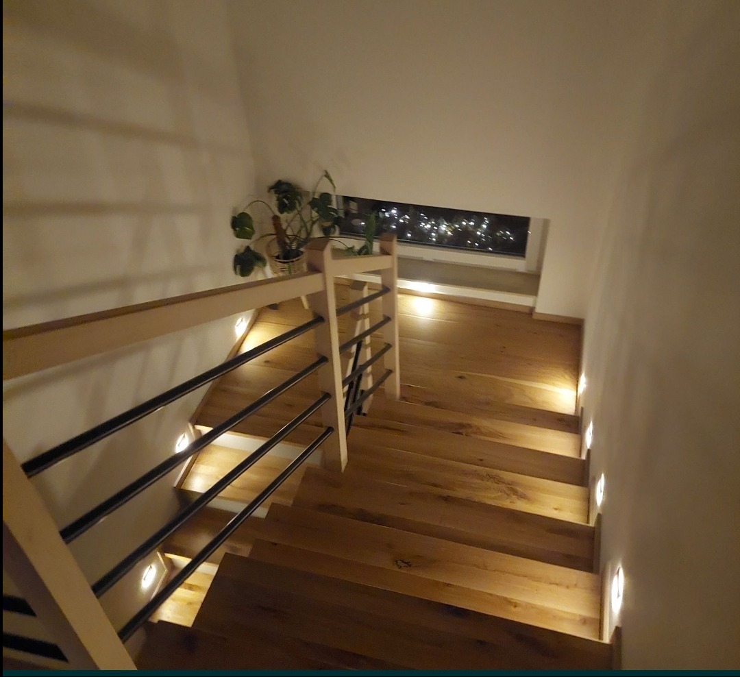 Drewniane schody z podświetleniem LED w stopniach i czarnymi, metalowymi balustradami, widok z góry, roślina doniczkowa na podeście.