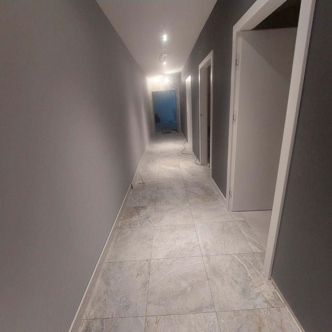 Długi korytarz z szarymi ścianami i podłogą wyłożoną płytkami imitującymi kamień. Białe drzwi po prawej stronie i widok na dalszą część korytarza z niebieskim zabezpieczeniem na końcu.