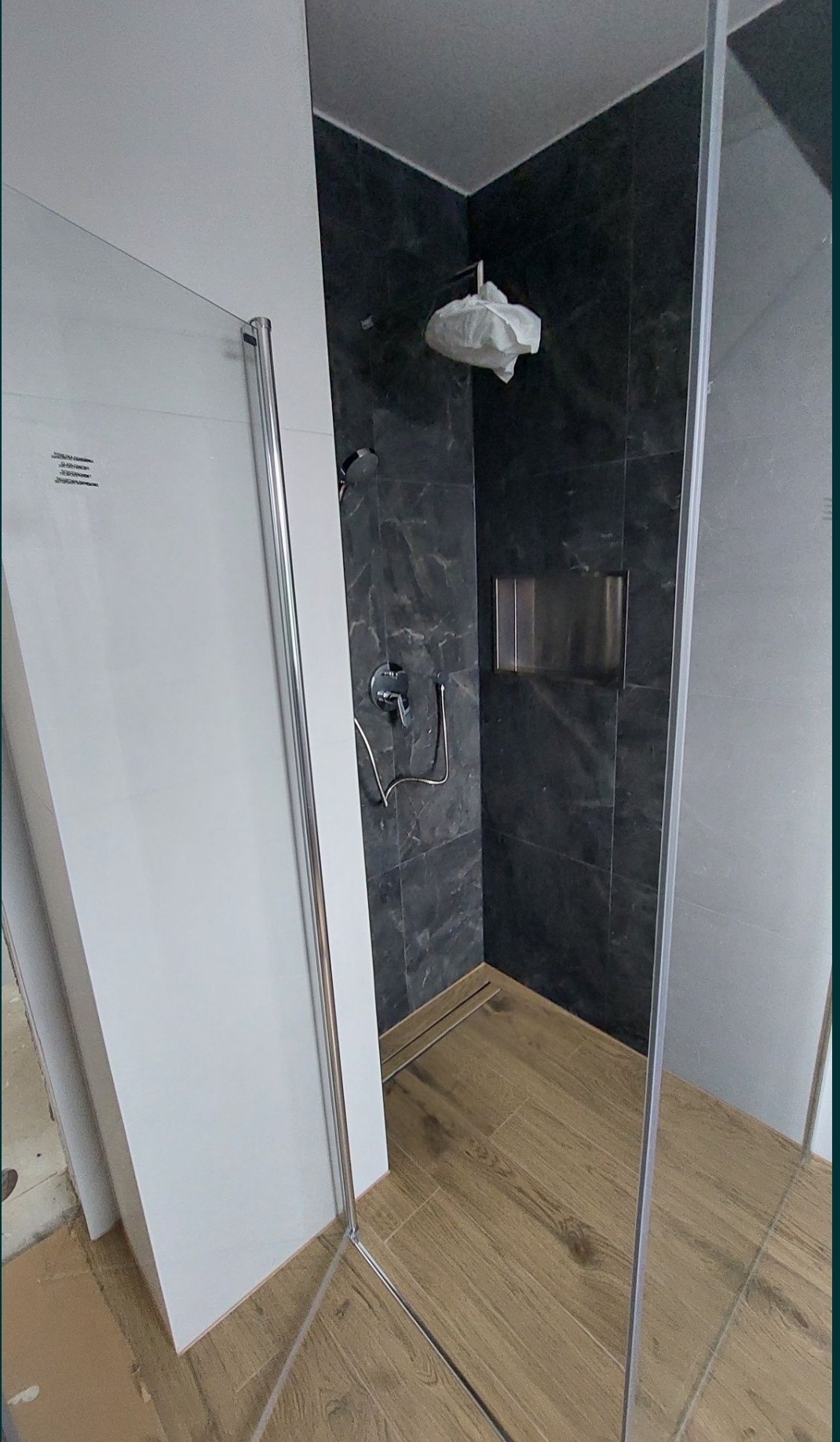 Nowoczesna kabina prysznicowa z czarną marmurową ścianą, chromowaną armaturą i drewnopodobną podłogą, z głęboką wnęką na kosmetyki w ścianie, oraz zabezpieczoną folią głowicą prysznicową.