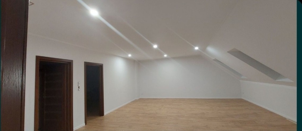 Poddasze po remoncie z jasną podłogą imitującą drewno, białymi ścianami, trzema wbudowanymi lampami sufitowymi i dwoma oknami dachowymi. Widoczne również dwie pary drzwi z ciemnego drewna.