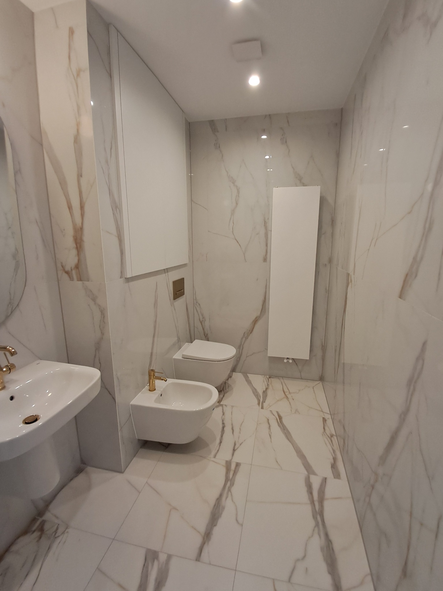 Elegancka łazienka wykończona marmurowymi płytkami z armaturą w kolorze złota, prezentująca umywalkę, bidet, toaletę i grzejnik.