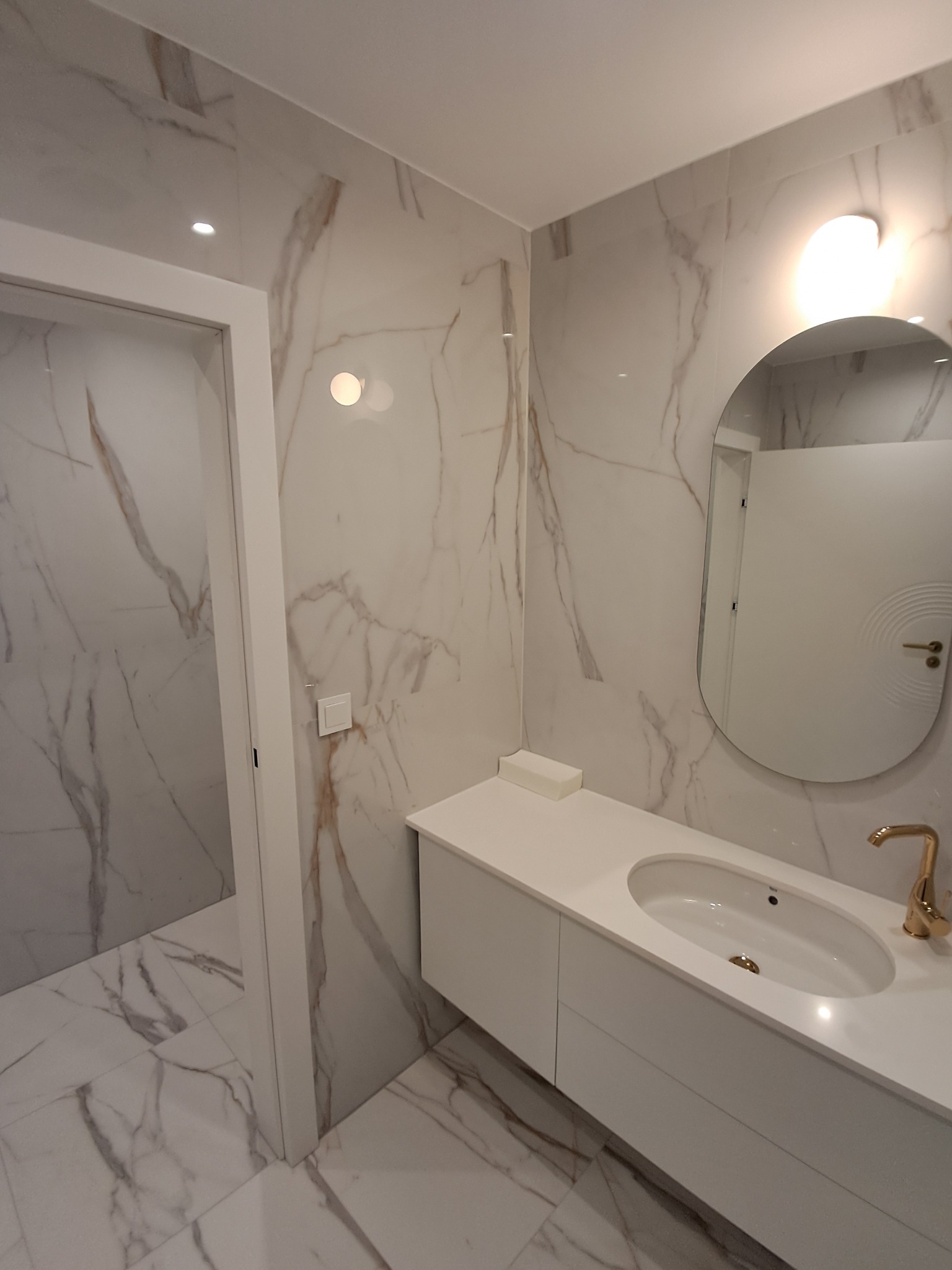 Nowoczesna łazienka z białymi marmurowymi płytkami na ścianach i podłodze, białą szafką pod umywalką z blatem i złotą baterią oraz owalnym lustrem z podświetleniem