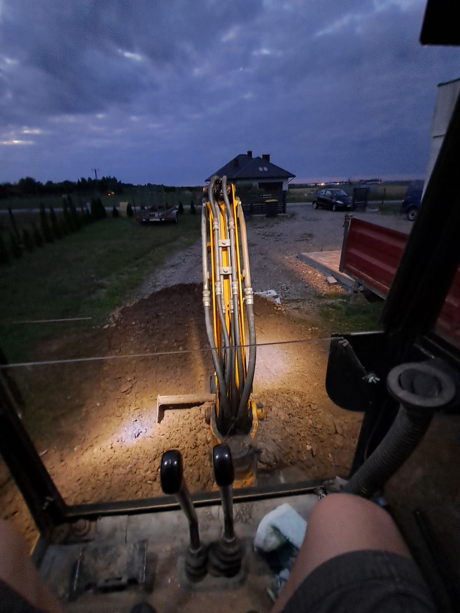 Widok z kabiny koparki na wykopany teren, w tle dom i pochmurne niebo o zmierzchu.