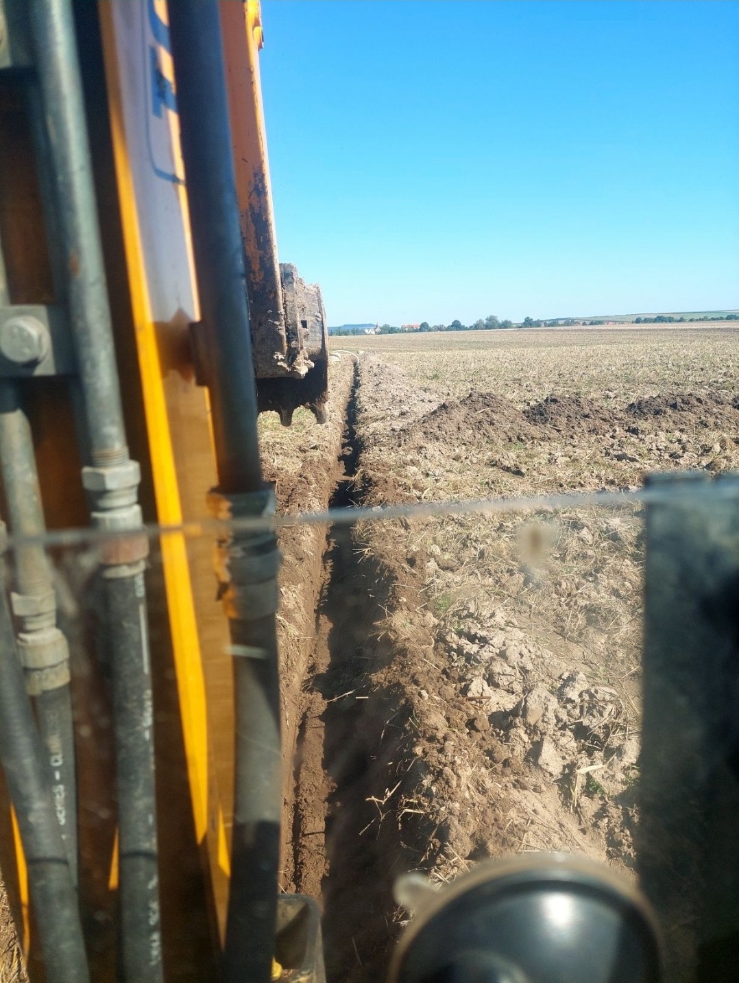 Wąski wykop w polu uprawnym widziany z kabiny koparki, z widocznymi przewodami hydraulicznymi i fragmentem lemiesza, błękitne niebo w tle.