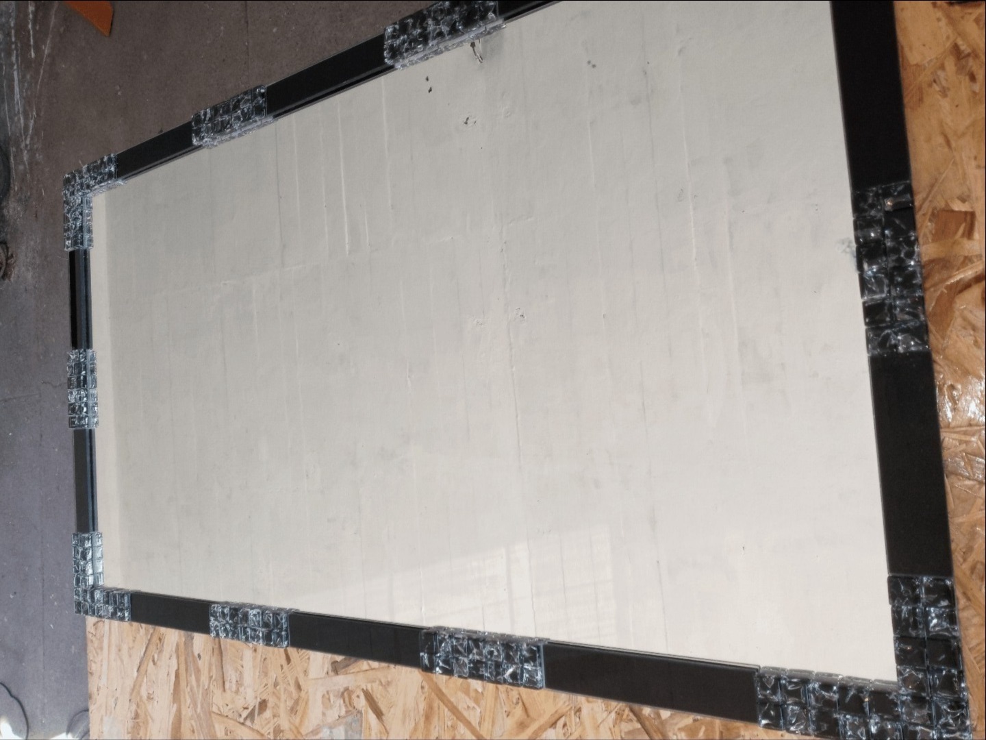 Prostokątne lustro z czarną ramą ozdobioną szklanymi kostkami, widoczne na tle płyty OSB.