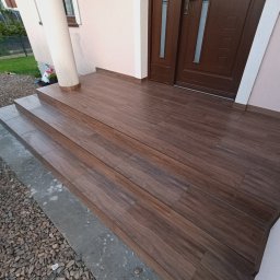 Marcin Skowronek ProDesign - Wykończone schody wejściowe do domu z brązowych płytek imitujących drewno, prowadzące do ciemnobrązowych drzwi wejściowych, z widoczną kolumną i fragmentem elewacji w tle.