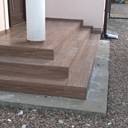 Marcin Skowronek ProDesign - Wykończone schody wejściowe do domu z brązowych płytek imitujących drewno, otoczone szarym żwirem, z widoczną białą kolumną i fragmentem zaparkowanego białego samochodu.