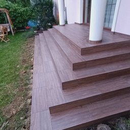 Marcin Skowronek ProDesign - Zewnętrzne schody i taras wyłożone brązowymi płytkami imitującymi drewno, z widocznymi słupami i fragmentem trawnika po bokach.