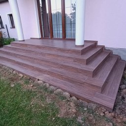 Marcin Skowronek ProDesign - Zewnętrzne schody wejściowe do domu wyłożone brązowymi płytkami imitującymi drewno, z widocznymi fugami, prowadzące do drzwi z kolumnami, otoczone trawnikiem i ziemią.