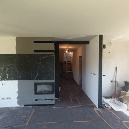 Marcin Skowronek ProDesign - Wnętrze w trakcie remontu: częściowo wykończony kominek z czarnym marmurem, fragment ściany z nałożoną gładzią, zabezpieczona podłoga, widoczne narzędzia i drabina.