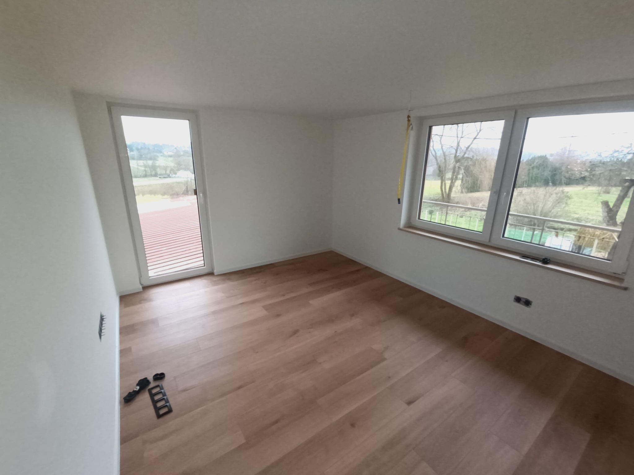 Pusty pokój po remoncie z nową podłogą z paneli, świeżo pomalowanymi ścianami na biało, widocznymi gniazdkami elektrycznymi i oknami z widokiem na zewnątrz. Na podłodze leżą narzędzia montażowe.